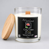 A 12 oz glass jar candle with white wax and a black label reading 'Rebecca's paradise Cozy Cabin with Ginger and Vanilla'. The candle is shown with its white lid resting on a brown cardboard box next to it.