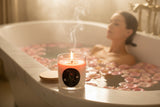 A clear glass jar candle with white wax and a pink wax top layer, shown with a light wood lid leaning against it on a white background. The front label reads 'Rebecca's paradise' and 'Rose Garden'.